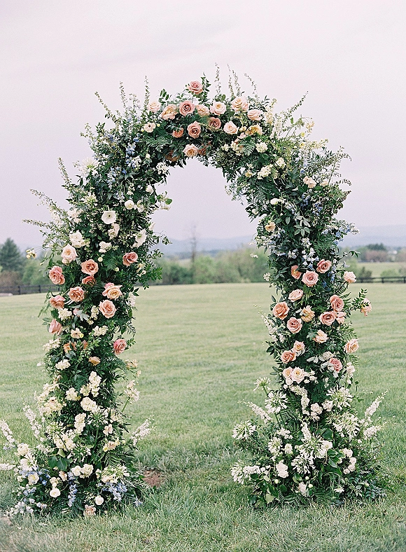 Floral ceremony arch with roses and white-blue blooms, greenery and ground flowers set on a grass lawn with hills and sky behind