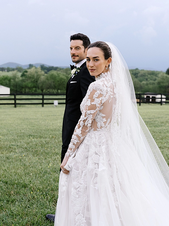 Couple portrait of bride looking back in lace dress and long veil beside groom in black tuxedo, in a grassy field by a wooden fence under clouds