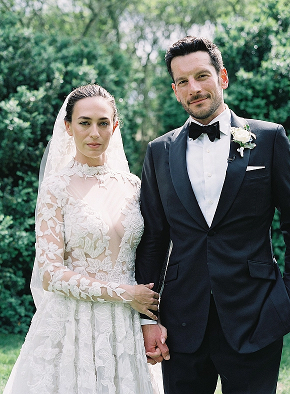 Couple portrait of bride in lace long-sleeve dress and veil holding hands with groom in black tuxedo, in sunlit garden greenery