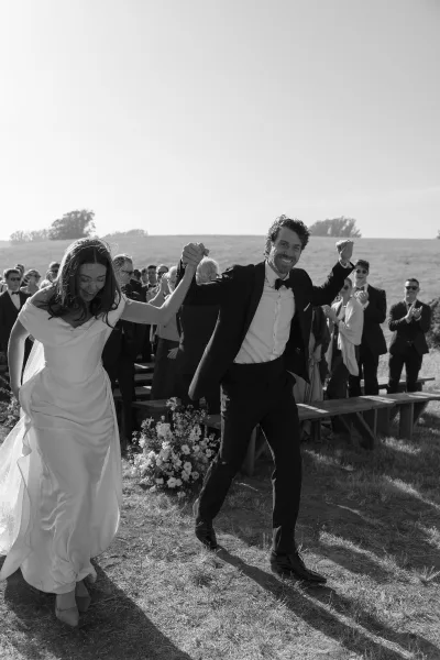 Wedding recessional as bride and groom walk hand in hand down a grassy aisle, guests clapping beside benches under open sky