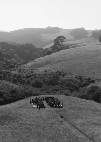Outdoor wedding ceremony with wooden benches and wedding guests arranged in rows on a meadow hilltop with rolling hills beyond
