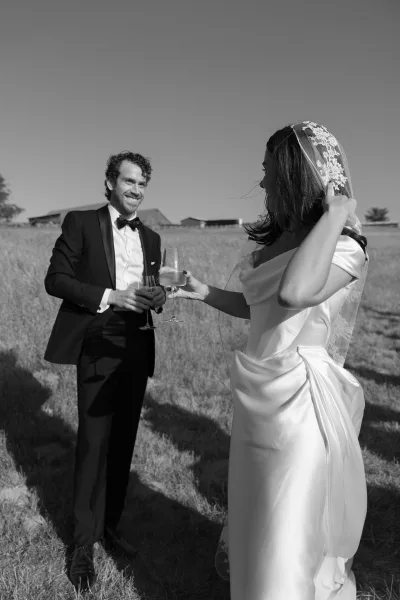 Wedding toast moment as bride and groom toast with champagne flutes, her lace veil and satin gown beside his black tuxedo in a grassy field
