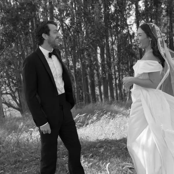Couple portrait of bride and groom laughing, her cathedral veil and off-the-shoulder dress beside his tuxedo in a woodland clearing