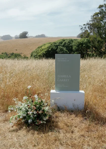 Wedding welcome sign on a white plinth with greenery and ground florals, set in a meadow with rolling hills and trees under open sky