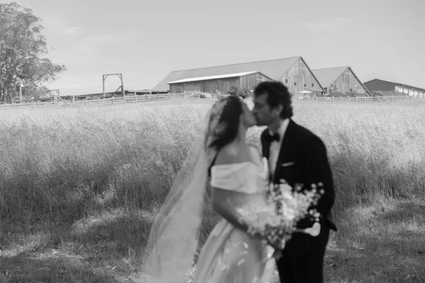Wedding kiss portrait of bride and groom kissing, her long veil flowing as they hold a bouquet in a meadow with barns behind