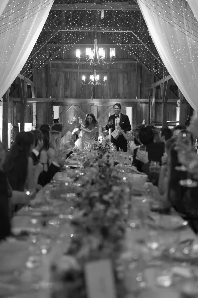 Wedding reception entrance as newlyweds raise champagne flutes under string lights and a chandelier in a barn filled with cheering guests