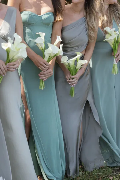 Bridesmaid dresses in sage green tones, mismatched satin gowns as bridesmaids hold white calla lily bouquets on a grass lawn outdoors