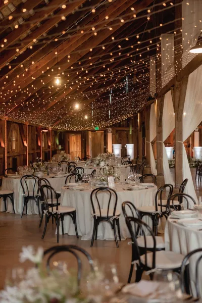 Reception tablescape with white linens and floral centerpieces on round tables under string lights in a rustic barn with wooden beams