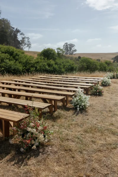 Ceremony seating with wood benches lines a meadow aisle, accented by white flowers and greenery, with hills, trees, and blue sky beyond