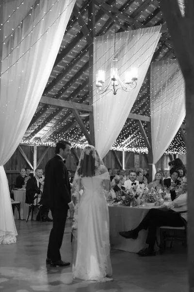 First dance under string lights as bride in wedding dress and veil twirls with groom in tuxedo in a rustic barn reception with candles