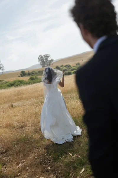 First look moment as groom in a black suit watches the bride from behind, her lace veil and long train flowing in a meadow of golden grass