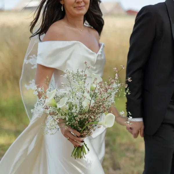 Couple portrait of bride and groom holding hands, bride with calla lily bouquet and veil, standing in tall grass field under open sky
