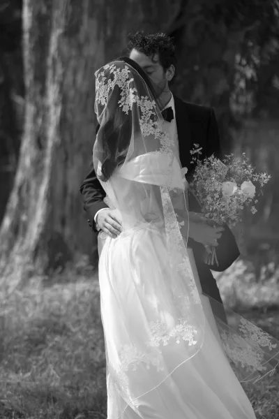 Wedding kiss portrait of bride and groom kissing under a lace veil, bride holding bouquet as groom in tuxedo stands in a garden setting