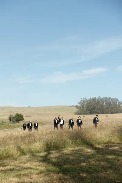 Groomsmen portrait of men in black tuxedos walking through tall grass in an open field, with rolling hills, trees, and blue sky behind them