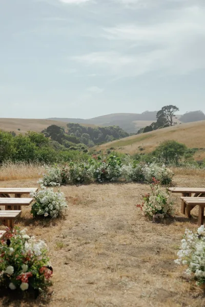 Ceremony aisle decor with white and green ground floral arrangements and wood benches set in a dry grass field under open sky