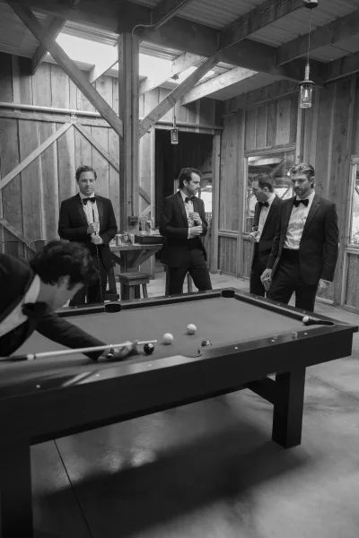 Groomsmen candid in tuxedos and bow ties playing pool, holding drinks around a table under pendant lights in a rustic barn bar setting
