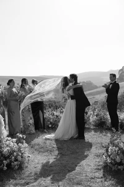 Wedding kiss as bride and groom embrace, long veil blowing behind them while wedding party watches in a meadow with rolling hills