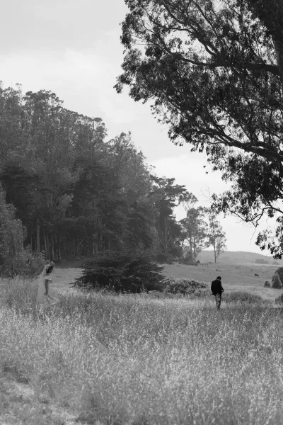 Couple portrait in a black and white wedding photo, bride in veil and groom in suit walking through a wildflower meadow toward hills and sky