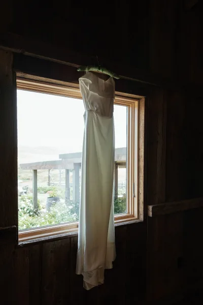 Wedding dress hanging by a window on a dress hanger, lit by natural light against a wood wall with porch and landscape beyond