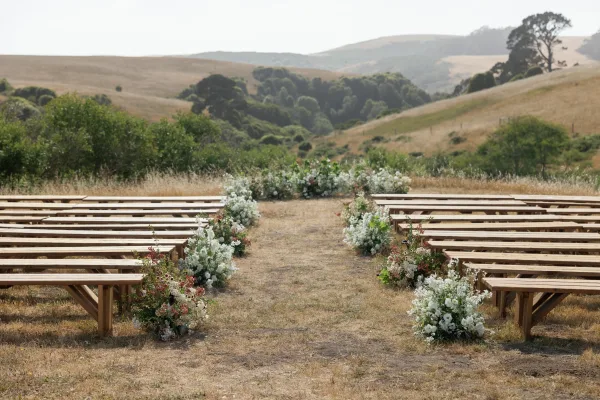Outdoor ceremony setup with a wedding ceremony aisle lined by white and green florals, wood benches, and meadow hills under open sky