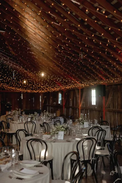 Reception tablescape with barn wedding reception vibe, round white linen tables, floral centerpieces, and café string lights under wood beams