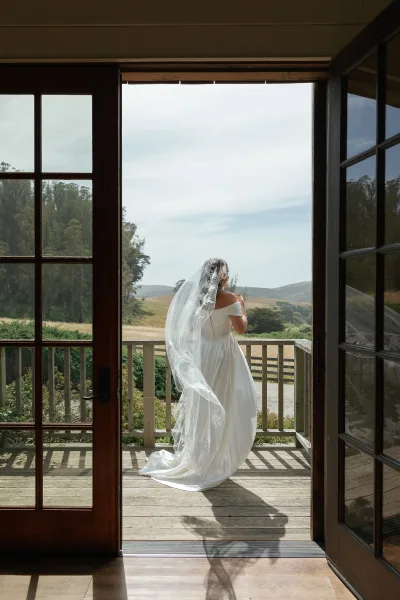 Bridal portrait of a bride from behind in an off the shoulder gown with a lace veil and long train at an open doorway overlooking hills