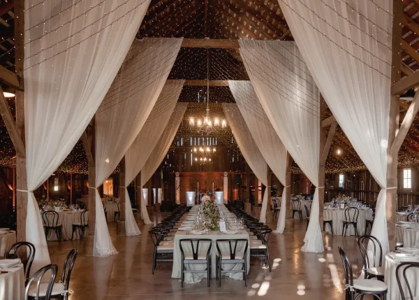 Wedding reception decor in a rustic barn wedding reception with white ceiling draping, string lights, banquet and round tables set for dinner