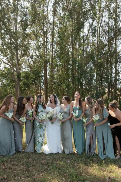 Bridesmaids group photo with bride and bridesmaids in sage green dresses holding calla lily bouquets in a sunny outdoor field with trees
