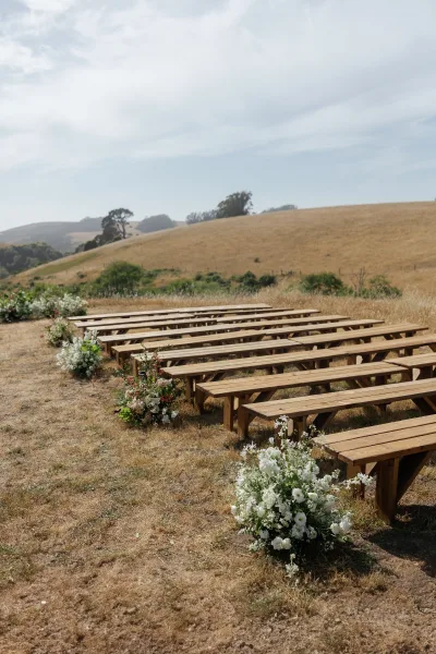 Outdoor ceremony seating with wedding bench seating, wooden benches lining an aisle with white flowers and greenery in a dry hillside field