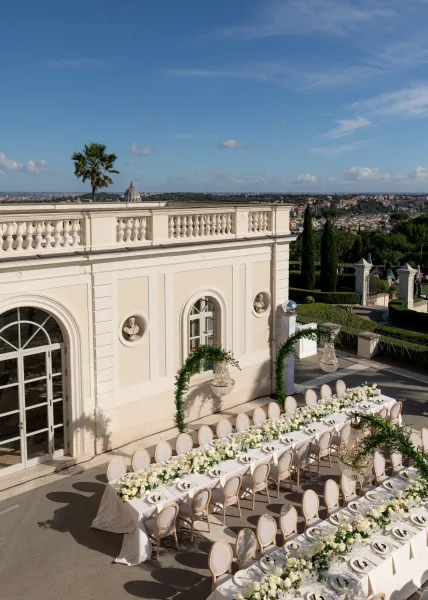 Reception tablescape with long banquet table wedding setup, white floral garlands and chandeliers on a villa terrace with skyline view