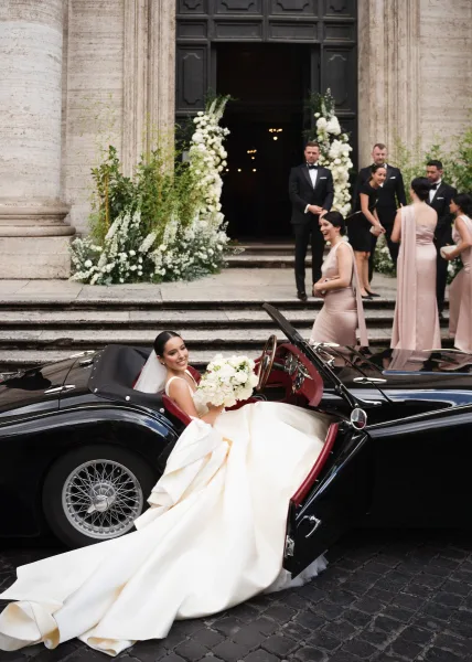 Bride in wedding car, a vintage wedding car with bouquet and veil as tuxedos and bridesmaids gather by a stone church doorway.