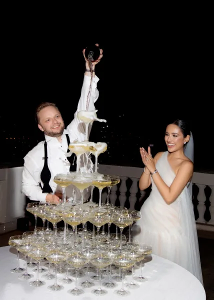 Champagne tower wedding display of coupe glasses as the groom pours from a bottle beside the bride on a balcony at night with distant lights
