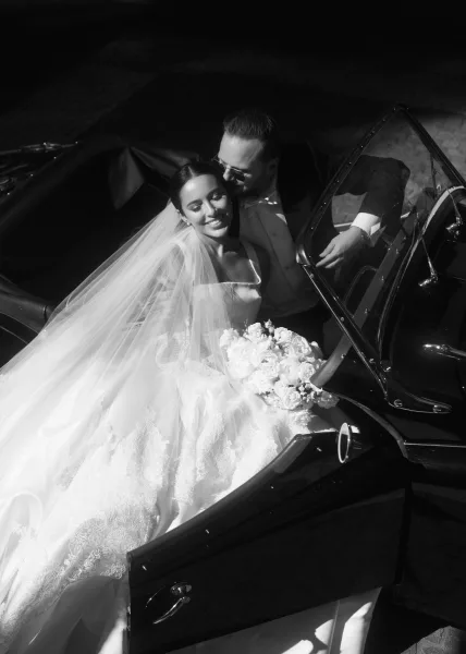 Couple portrait of newlyweds in a vintage convertible car, bride in lace dress with veil and bouquet, groom in tux and sunglasses