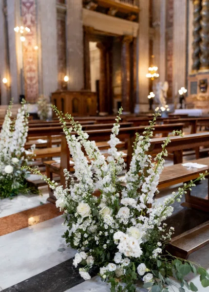 Ceremony aisle decor with church wedding aisle flowers, white florals and greenery lining wooden pews on a marble floor toward an ornate altar