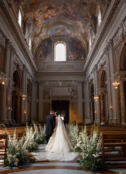 Wedding kiss portrait of bride and groom kissing in a grand church aisle, her cathedral veil trailing past white florals and pews