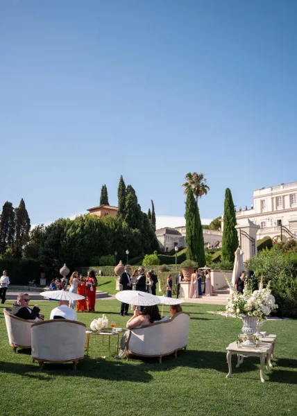 Wedding cocktail hour with guests mingling in formalwear around outdoor lounge seating under white parasols on a manicured villa lawn