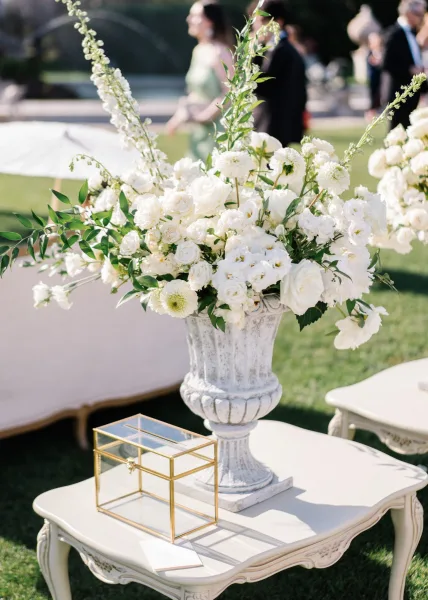 Wedding floral arrangement of white wedding flowers with roses and greenery in a stone urn on a vintage table, blurred guests behind