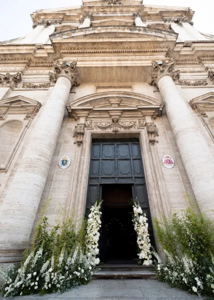 Ceremony entrance decor with white florals on tall pillars and greenery ground arrangements framing a carved stone church doorway and steps