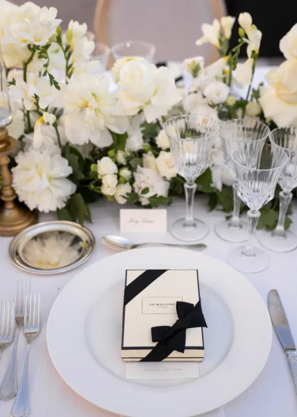 Wedding tablescape with a white wedding centerpiece and greenery, crystal wine glasses, black ribbon details, and gold candlestick on white linens