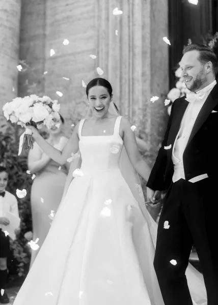 Wedding recessional as bride and groom walk out smiling, holding hands in a rose petal toss at stone-columned entrance with guests cheering