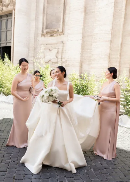 Bride with bridesmaids helping bride hold a white rose bouquet and dress train, with veil and blush dresses in a stone courtyard.