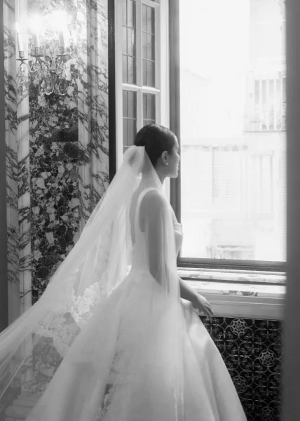 Bridal portrait of a bride looking out window in side profile, wearing a strapless gown and long veil train by a marble wall sconce