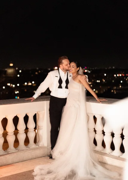 Wedding couple portrait of bride and groom on balcony as he kisses her forehead, leaning on railing with veil and city lights behind