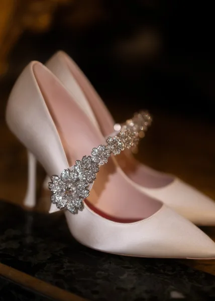 Wedding shoes with bridal heels in satin, detailed with a rhinestone shoe clip, resting on a dark surface with soft blur behind
