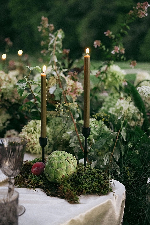 Reception centerpiece with taper candle centerpiece on a moss table runner, featuring artichoke, fruit, and hydrangea in garden greenery