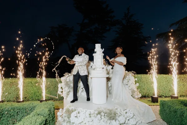 Champagne pop moment as the groom popping champagne sprays beside sparkler fountains, set against garden hedges and trees under the night sky.
