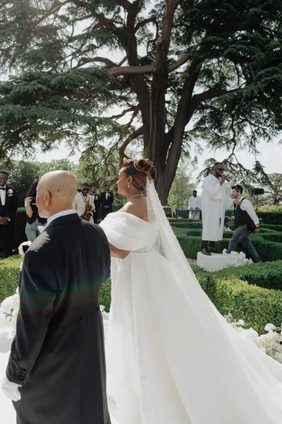 Ceremony processional with a bride walking down aisle, her long veil trailing along a white aisle beneath a large tree and garden hedges.