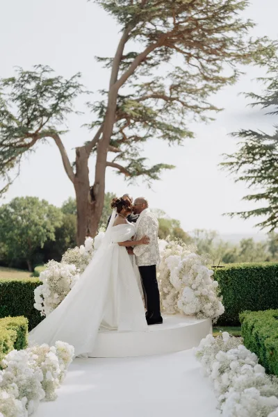 Ceremony kiss as the bride and groom embrace at an outdoor garden ceremony, framed by abundant white florals among hedges and lawn.