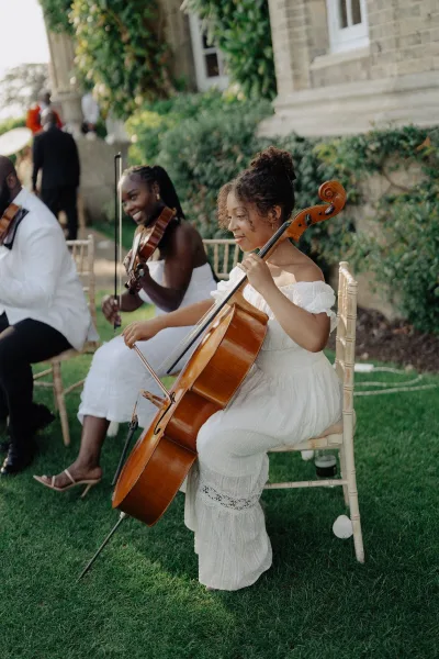 Wedding musicians deliver a string ensemble wedding performance on a garden lawn by an ivy-covered stone facade, featuring a cello.