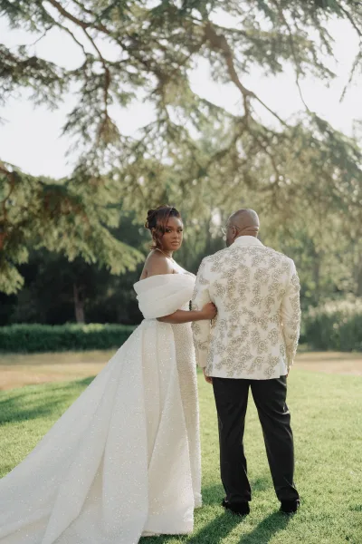 Couple portrait with the bride holding the groom's arm, her long train trailing across a sunlit garden lawn under garden trees.